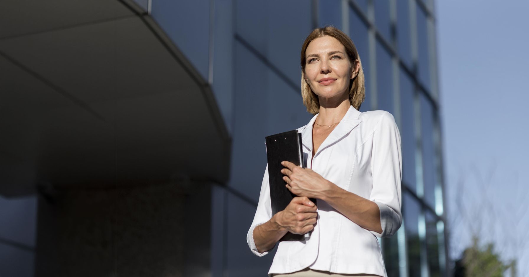 A professional woman in a white blazer holding a folder, standing confidently outside a modern glass office building.