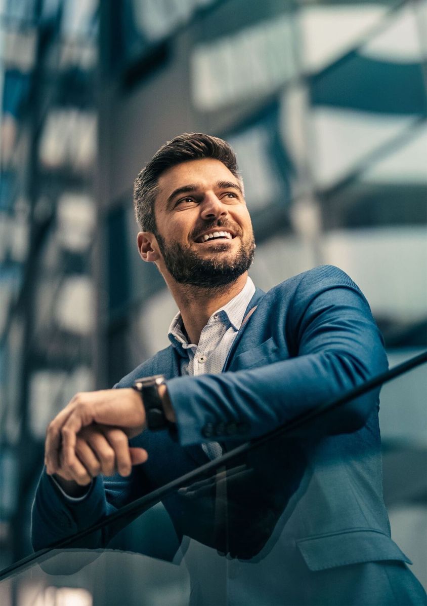 A smiling businessman in a blue suit leaning on a glass railing, surrounded by modern glass architecture.